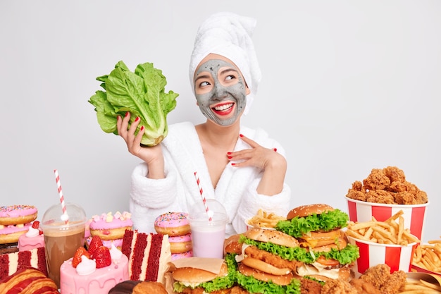 Cheerful dreamy young woman undergoes beauty procedures at home looks happily away holds green romaine lettuce