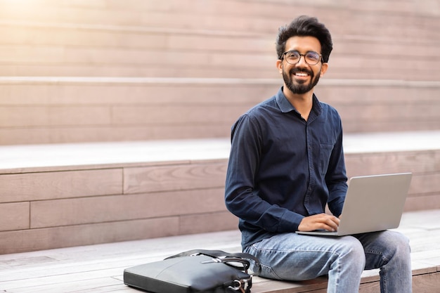 Cheerful eastern guy freelancer sitting on stairs using laptop