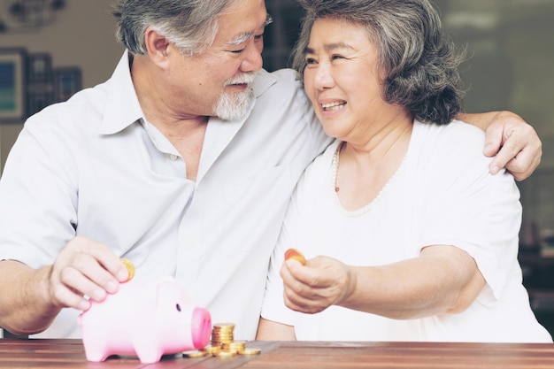사진 명랑한 노인 부부는 돼지 저금통에 동전을 넣고 나무 탁자에 동전을 쌓아 돈을 절약하며 함께 행복하게 웃고 있습니다