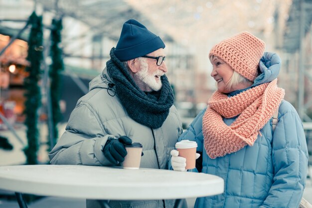 屋外で一緒に立って、円卓にコーヒーのカートンカップを置く暖かいコートを着た陽気な老夫婦