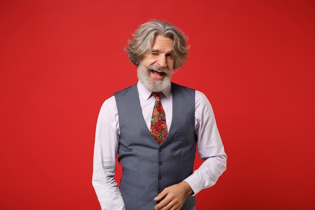 Cheerful elderly gray-haired mustache bearded man in classic shirt vest and colorful tie posing isolated on red wall background studio portrait. People lifestyle concept. Mock up copy space. Blinking.