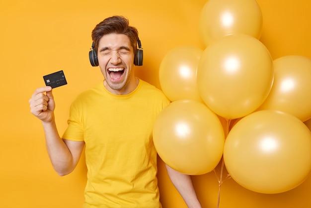 Cheerful emotional adult man exclaims loudly glad to get salary holds banking card holds bunch of inflared balloons ready for celebration dressed casually isolated over vivid yellow background