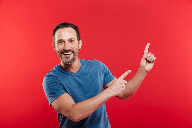 Cheerful emotions of unshaved man 30s in casual blue t-shirt smiling and pointing fingers upward on copyspace closeup, isolated over red background