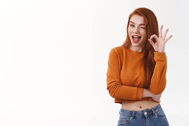 Cheerful enthusiastic redhead silly cheeky woman wink cunning show okay ok deal gesture smiling delighted hinting assure secret safe stand amused white wall give approval great choice