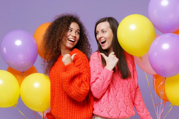 Photo cheerful european african american women friends in knitted sweaters isolated on violet purple background. birthday holiday party concept. celebrating hold air balloons pointing thumbs at each other.