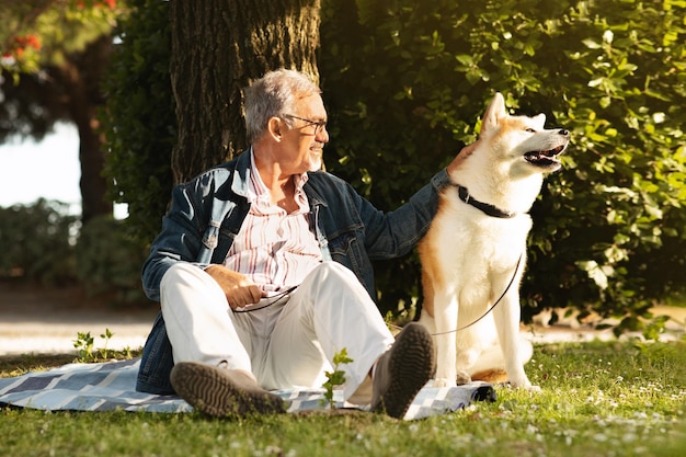 カジュアルなひげを生やした陽気なヨーロッパの老人は、犬と一緒に散歩を楽しみ、草の上に座ってリラックスします