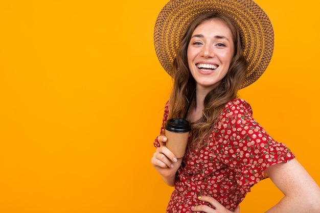 Cheerful european woman laughs on an orange background, a girl in a hat and a red dress with coffee in hand