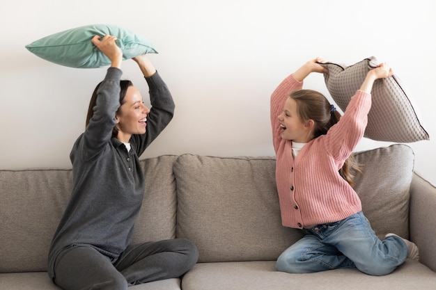 Cheerful excited european millennial mother and little girl fight pillow have fun together in living