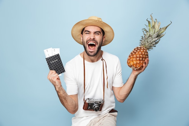 Cheerful excited man wearing blank t-shirt standing isolated over blue wall, holding pineapple, showing passport