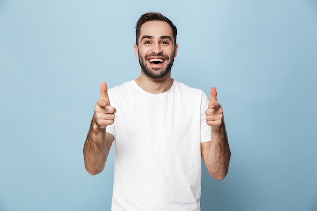 青い壁の上に孤立して立って、ポインティング空白のTシャツを着て陽気な興奮した男