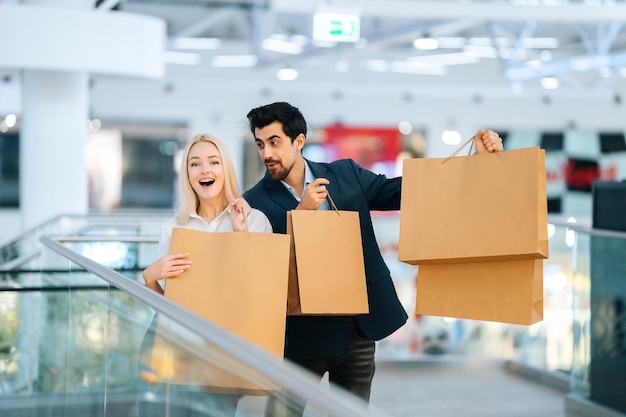 Cheerful excited young couple holding showing shopping bags with purchases and looking at camera in