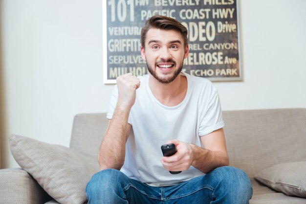 Cheerful excited young man using remote control and watching tv on sofa