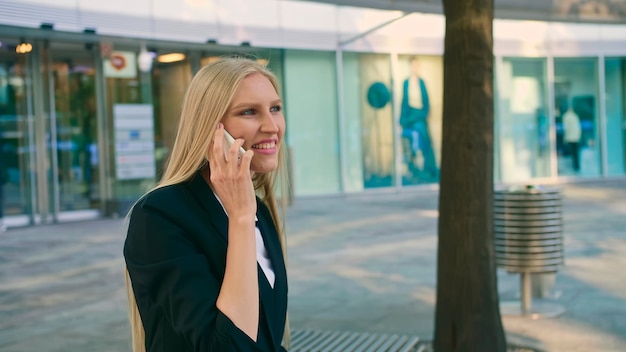 Cheerful executive girl waving with hand. Adult blond businesswoman in suit sitting on bench and waving with hand greeting colleague on street.