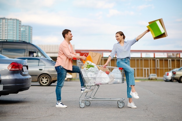 Cheerful family couple with bags in cart on supermarket car parking. Happy customers carrying purchases from the shopping center, vehicles