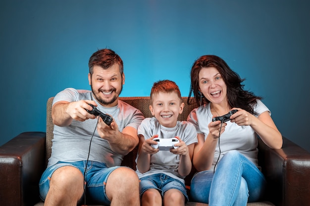 A cheerful family, dad mom and son play on the console, video games, emotionally react sitting on the couch. Day off, entertainment, leisure, spend time together.