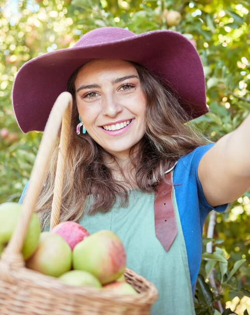 Cheerful farmer harvesting juicy organic fruit in season to eat Portrait of a happy woman taking selfies while holding basket of fresh picked apples on sustainable orchard farm outside on sunny day
