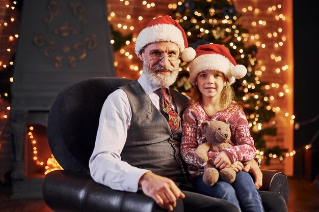 Cheerful fashioned senior man with grey hair and beard sitting with little girl in christmas hats and teddy bear.