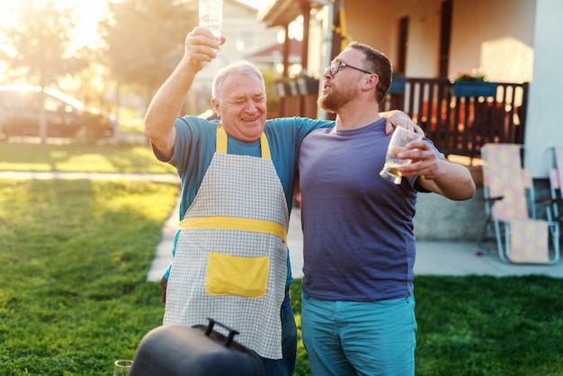 Suocero allegro e genero che abbracciano e che bevono birra mentre stando accanto alla griglia in cortile. concetto di riunione di famiglia.