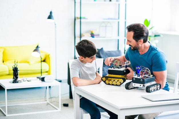 Cheerful father and son sitting at the table while constructing their robotic devices