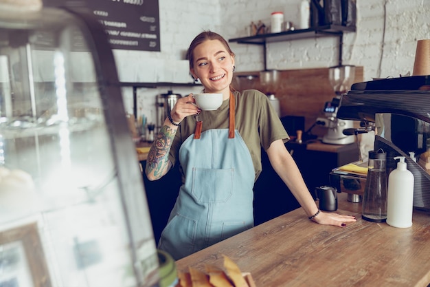 Cheerful female barista drinking coffee in cafeteria