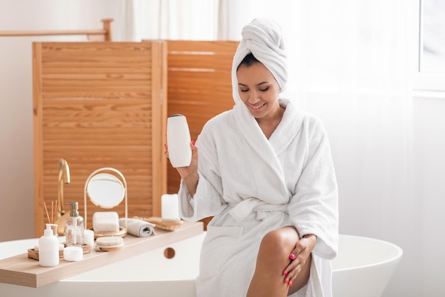 Cheerful female in bathrobe showing shampoo bottle in modern bathroom