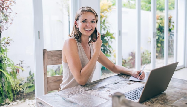 Cheerful female busy with freelance work from home using laptop taking notes making phone call