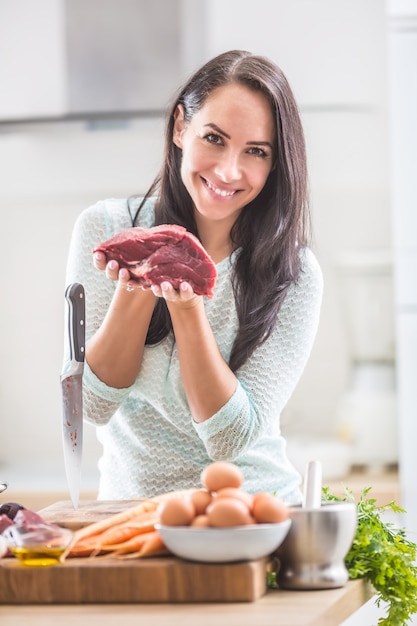 Cheerful female cook in kitchen holding raw beef meat.