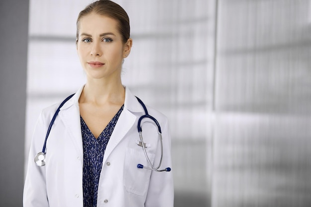 Cheerful female doctor standing in clinic. Portrait of friendly smiling woman physician. Perfect medical service in hospital. Medicine concept.