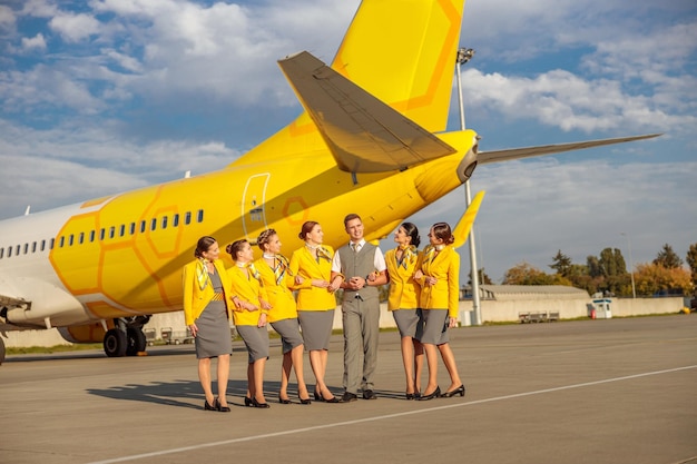 Cheerful female flight attendants looking at male colleague and smiling while standing near passenger airplane at airport