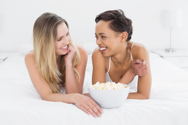 Cheerful female friends with popcorn bowl lying in bed
