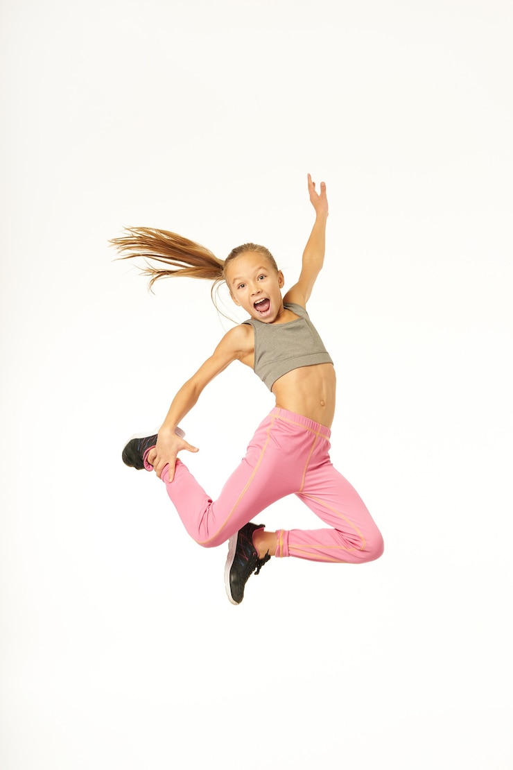 Photo cheerful female kid in sportswear jumping in the air and placing hand on her leg. isolated on white background