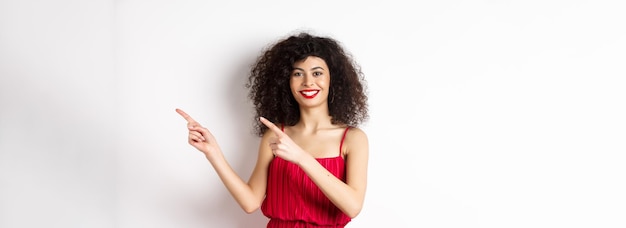 Cheerful female model in fashionable red dress smiling and pointing fingers right at logo standing o
