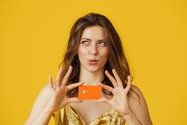 Cheerful female shopaholic showing credit card looking aside and thinking isolated on yellow