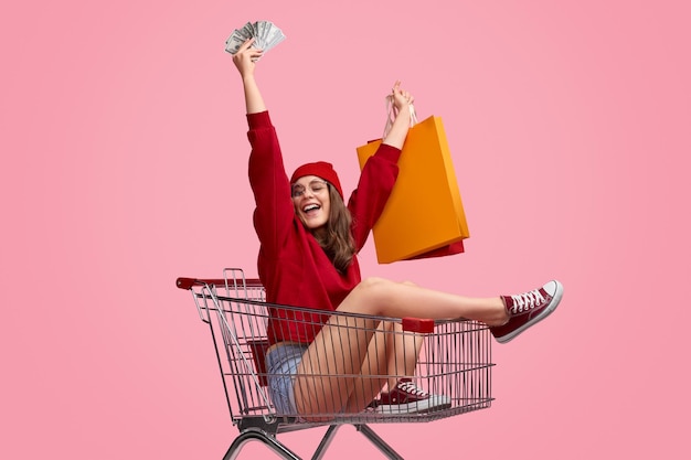 Cheerful female with money and shopping bags in trolley