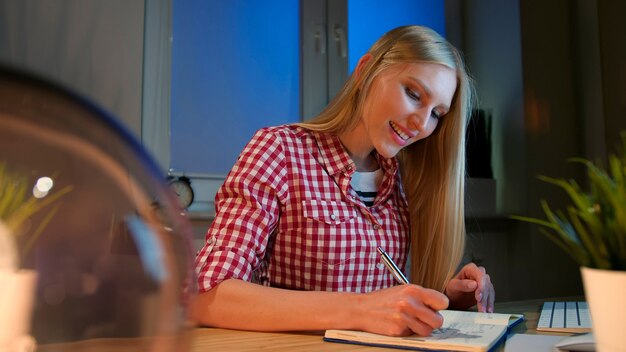 Cheerful female writing in daily planner lively young blond woman in casual checkered shirt sitting at wooden desk with notebook holding metal pen and looking at camera smiling at night in dark room