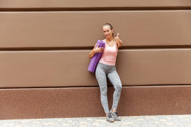 Photo cheerful fit woman with yoga at posing near the wall outdoor