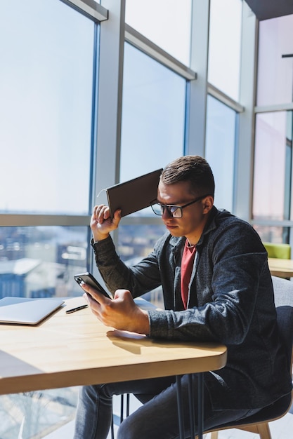 Cheerful freelancer in glasses browsing smartphone and smiling while sitting at table with laptop and notepad in cafe during daytime Working remotely from the office