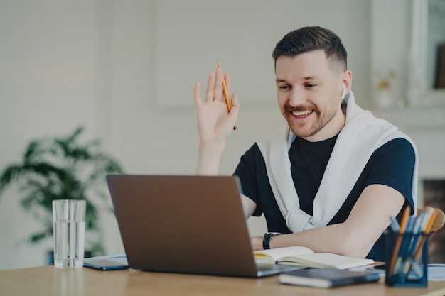 Cheerful freelancerbusinessman in earphones waves greets colleaguesclients in video conference from home office