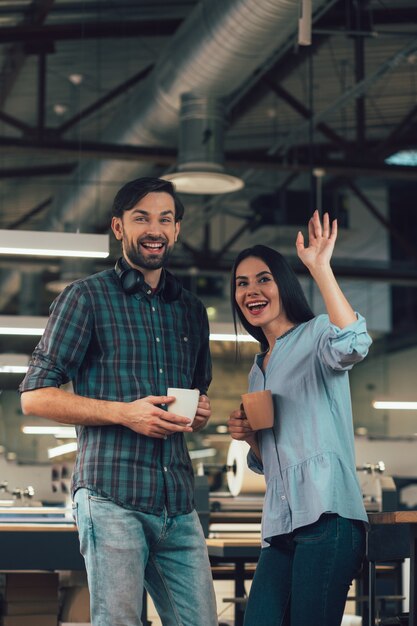 Cheerful friendly man and woman standing in the office with mugs and smiling. Happy lady waving to someone