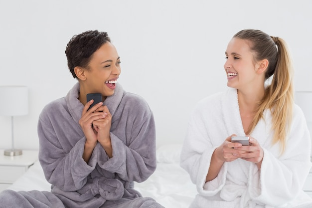 Cheerful friends in bathrobes text messaging on bed