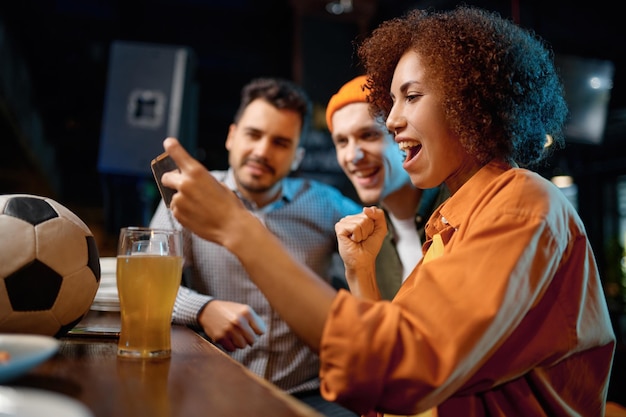 Фото Веселые друзья весело смотрят футбольный матч на смартфоне в спорт-баре. чрезвычайно эмоциональные молодые люди пьют разливное пиво за барной стойкой в пабе