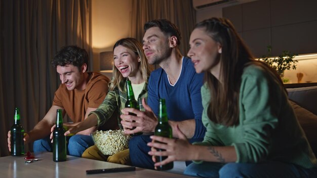 Foto amici allegri che guardano la televisione di notte, persone che bevono birra.