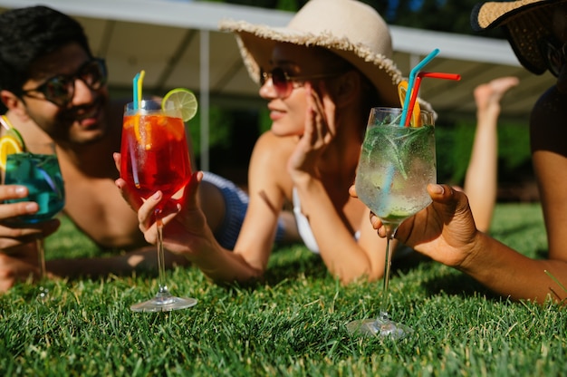 Cheerful friends with coctails rest on the grass near the pool. Happy people having fun on summer vacations, holiday party at the poolside outdoors. One man and two women are sunbathing