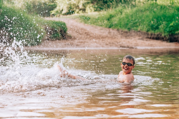 Cheerful funny boy 7-10 lies in river wearing goggles for swimming and splashes in rays of sun