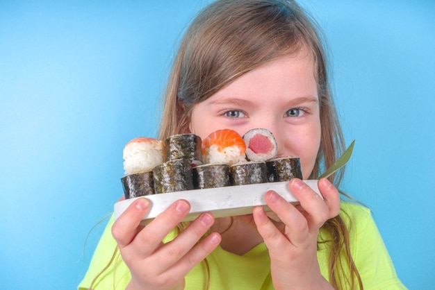 Cheerful funny caucasian blond preschool child girl with various sushi roll and chopsticks. On bright blue wall