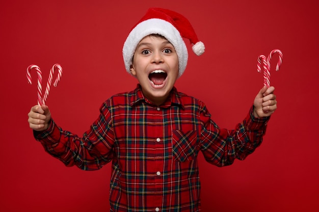 쾌활하고 재미있는 소년, 아름다운 꼬마는 크리스마스 사탕 지팡이를 들고 색색의 빨간색 배경에 포즈를 취하고, 손에 달콤한 줄무늬 막대 사탕을 들고, 카메라를 보고 기뻐합니다. 복사 공간이 있는 새해 개념