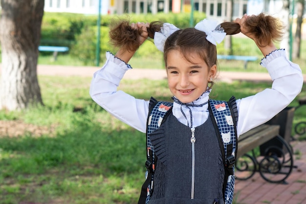 Cheerful funny girl with a toothless smile in a school uniform with white bows in school yard Back to school September 1 Happy pupil with a backpack Primary education elementary class