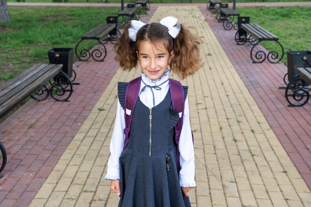 Cheerful funny girl with a toothless smile in a school uniform with white bows in school yard Back to school September 1 Happy pupil with a backpack Primary education elementary class