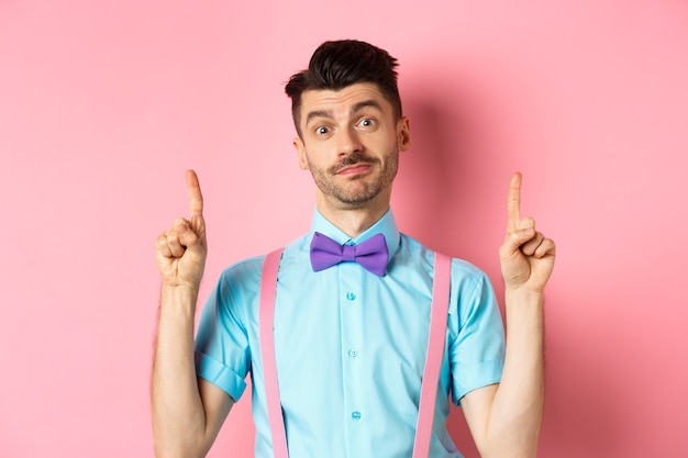 Cheerful funny man in bow-tie pointing fingers up, showing advertisement and smiling indecisive, standing over pink. Copy space