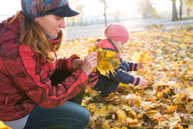 Cheerful and funny mother plays with her positive daughter yellow maple leaves while walking in a sunny autumn park. Concept of good family traditions